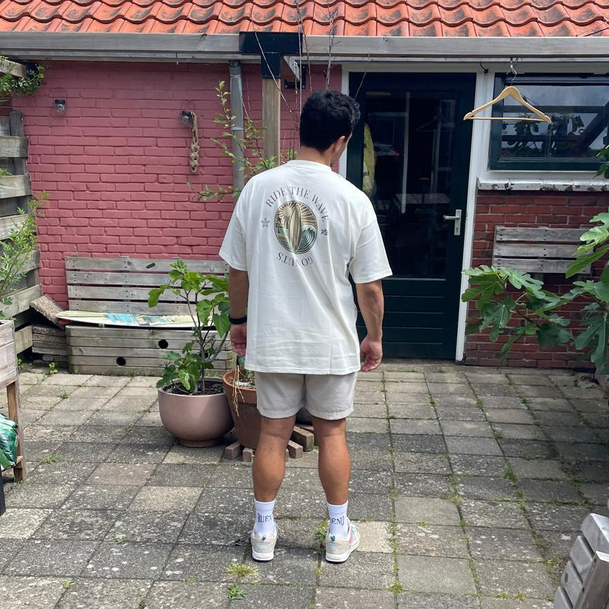 A man viewed from behind wearing a the Bueno Go Nuts Ride the Wave shirt with beige shorts, and white sneakers, standing on a paved backyard patio in front of a pink brick house. 
